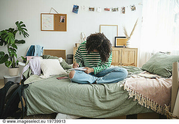Teenage girl writing in diary and sitting on messy bed at home