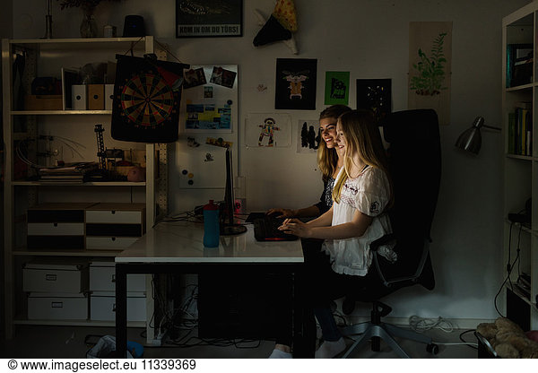 Teenage girls using computer at table in darkroom at home