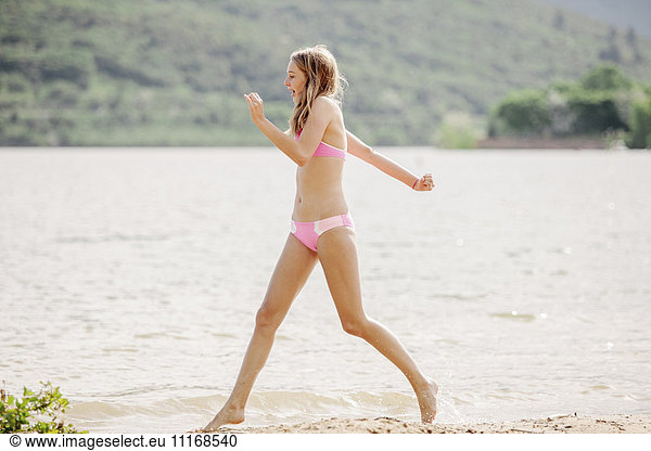 Teenage girls wearing a pink bikini running along a sandy beach by a lake.