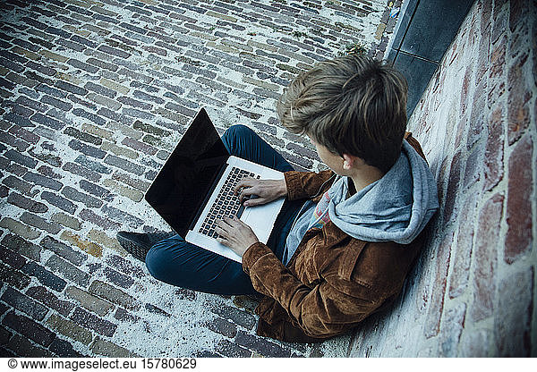 Teenager  der einen Laptop benutzt und auf einem Steinboden in der Stadt sitzt