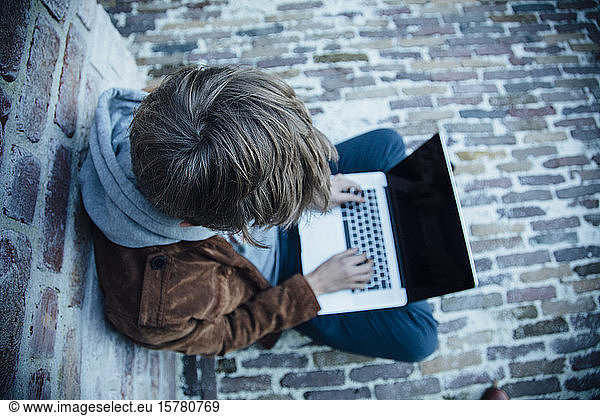 Teenager  der einen Laptop benutzt und auf einem Steinboden in der Stadt sitzt