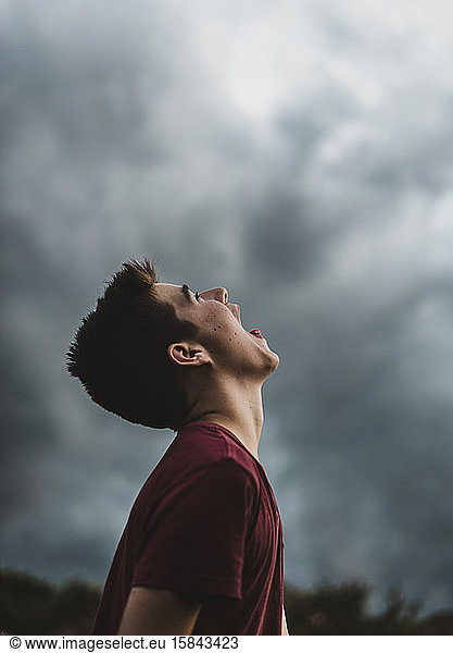 Teenager-Junge  der mit weit geöffnetem Mund in den dunklen Wolkenhimmel schaut.