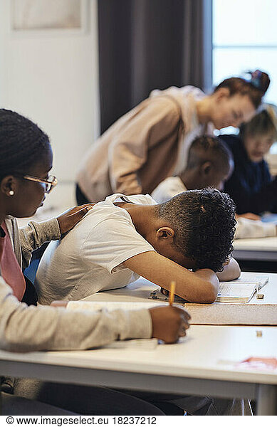 Teenager-Mädchen  das einen deprimierten männlichen Schüler tröstet  der seinen Kopf im Klassenzimmer auf den Schreibtisch legt
