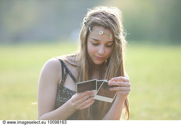 Teenager-Mädchen mit Gänseblümchen-Kopfbedeckung beim Betrachten von Sofortbildern im Park