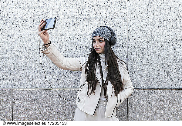 Teenager-Mädchen mit langen Haaren hören Musik durch Kopfhörer während der Einnahme selfie gegen Silber Wand