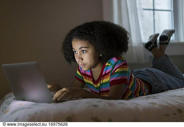 Ten year-old bi-racial girl working on her apple laptop lying on bed