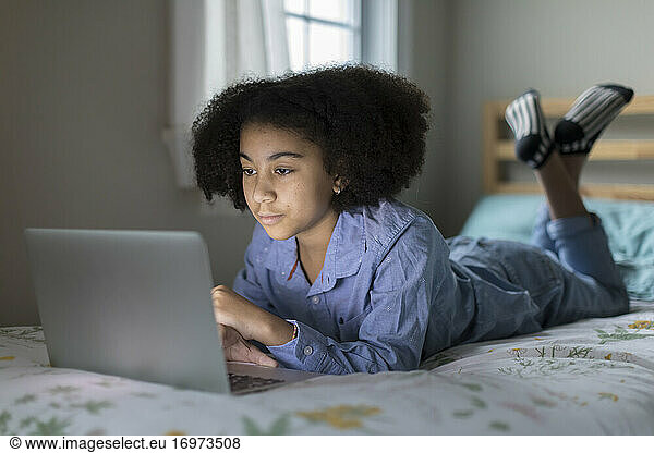 Ten year-old bi-racial girl works on her laptop while lying on bed