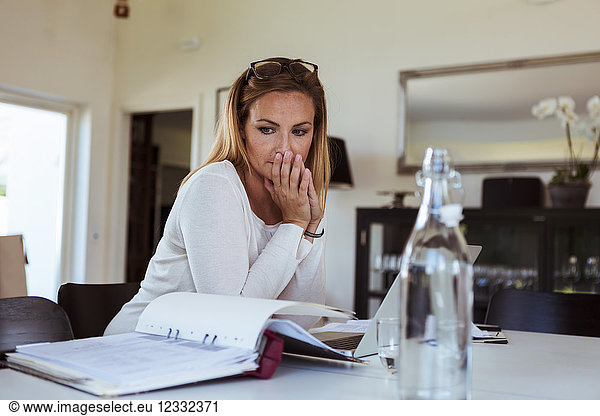 Tensed woman looking at bills while sitting at table in living room