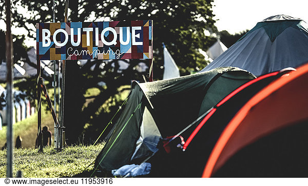 Tents on grass in a camping area at an outdoor music festival in summer. Sign reading Boutique Camping. Glamping.