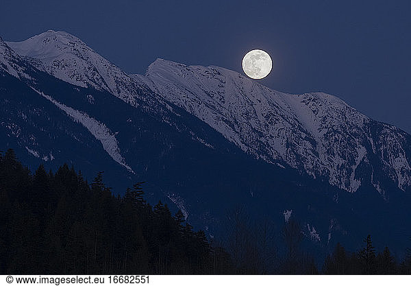 The full moon rises above snow covered mountain peaks on a spring night in the Coast Mountains of British Columbia.