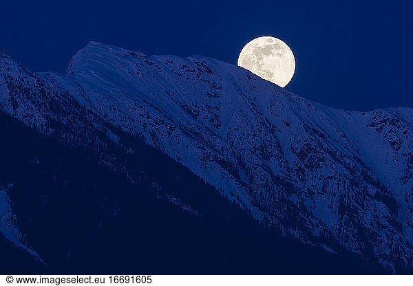 The full moon rises above snow covered mountain peaks on a spring night in the Coast Mountains of British Columbia.