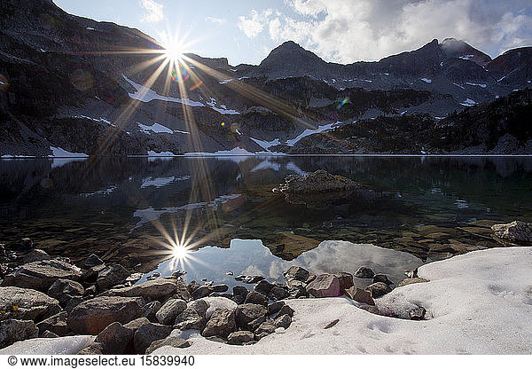 The sun sets over an alpine lake surrounded by mountain peaks.