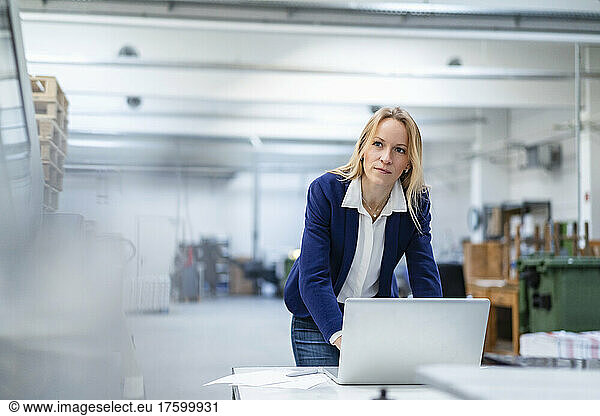 Thoughtful blond businesswoman with laptop leaning on desk in factory