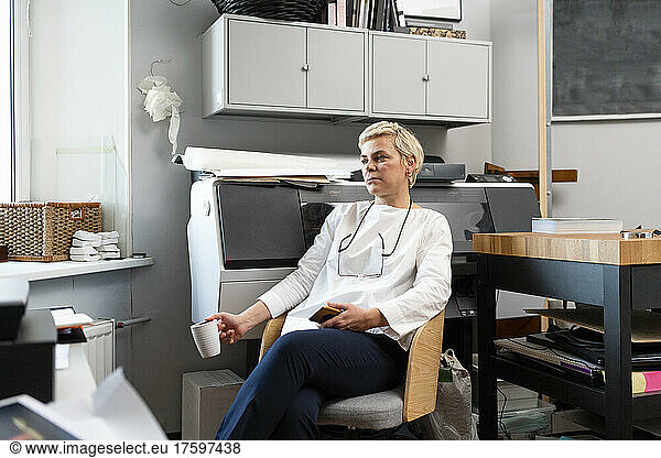 Thoughtful blond woman holding mug sitting on chair at workplace