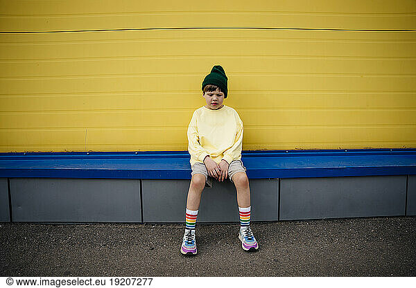 Thoughtful boy wearing sweatshirt sitting in front of yellow wall