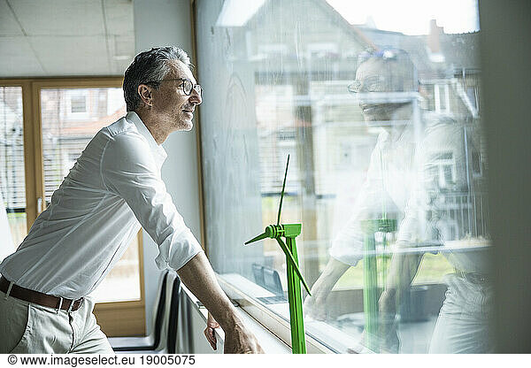 Thoughtful businessman by wind turbine model looking out through window at office