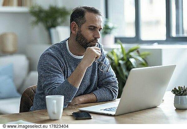 Thoughtful businessman holding eyeglasses looking at laptop working from home
