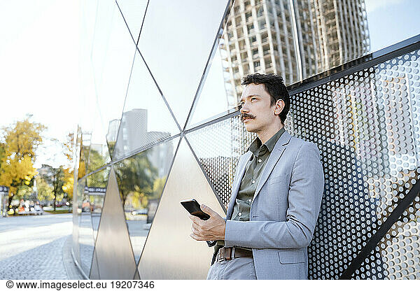 Thoughtful businessman holding smart phone in front of modern glass building