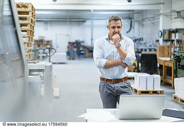 Thoughtful businessman looking at laptop working in factory