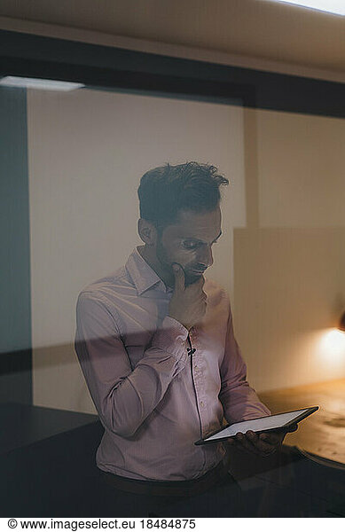 Thoughtful businessman using tablet PC seen through glass in office