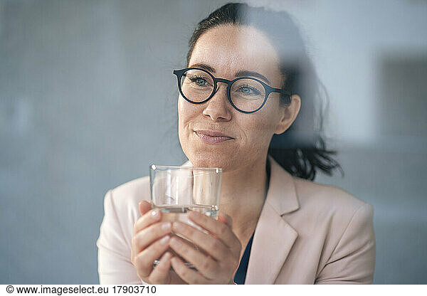 Thoughtful businesswoman holding drinking glass in front of wall