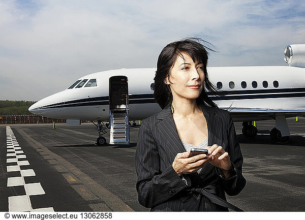 Thoughtful businesswoman holding phone while standing against corporate jet on runway
