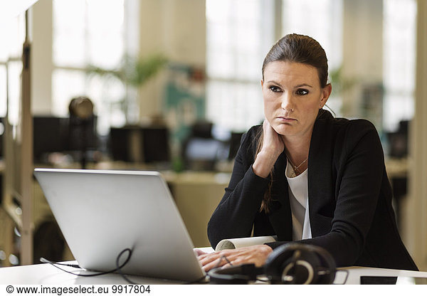 Thoughtful businesswoman looking away while using laptop at desk in creative office