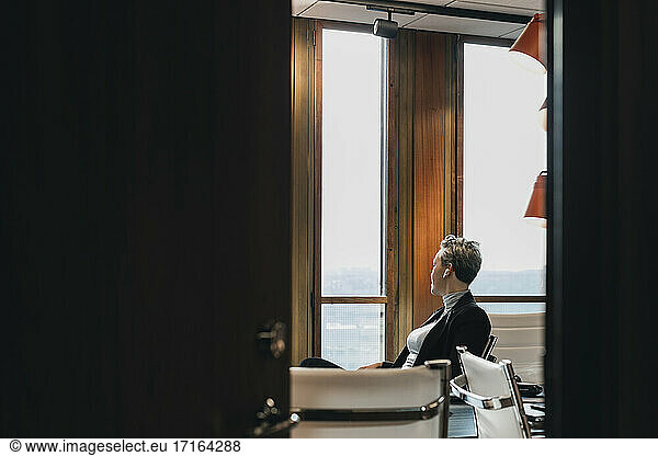 Thoughtful businesswoman looking through window sitting in board room seen from doorway