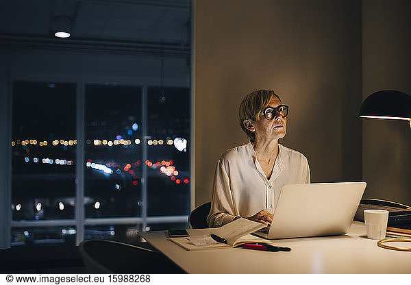 Thoughtful businesswoman sitting with laptop at illuminated desk in creative office
