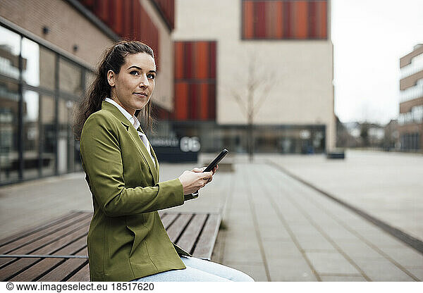 Thoughtful businesswoman sitting with mobile phone on bench outside building