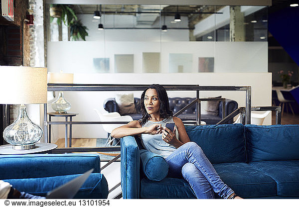Thoughtful businesswoman using mobile phone while sitting on sofa at creative office