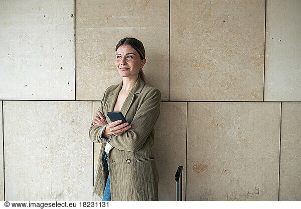 Thoughtful businesswoman with arms crossed holding smart phone in front of wall