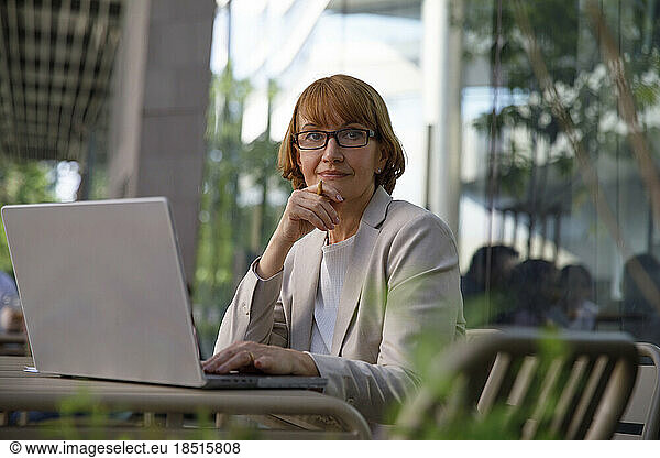 Thoughtful businesswoman with hand on chin sitting at sidewalk cafe with laptop