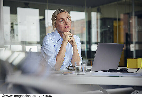 Thoughtful businesswoman with hand on chin sitting by laptop at desk in office