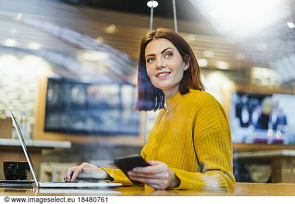 Thoughtful businesswoman with smart phone and laptop sitting at table in cafe
