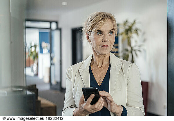 Thoughtful businesswoman with smart phone in office seen through glass