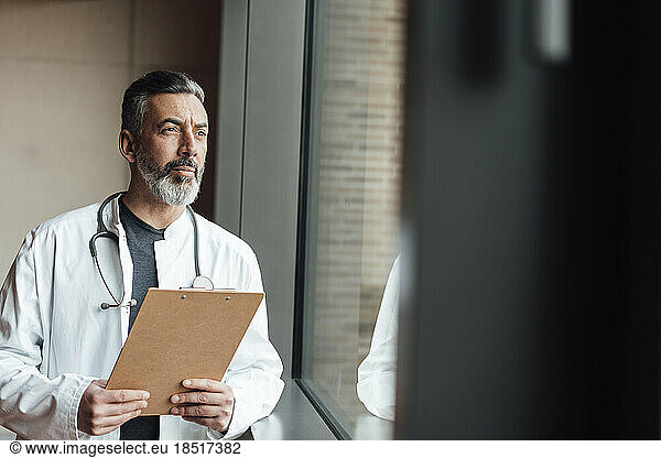 Thoughtful doctor standing with clipboard by window