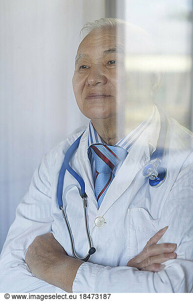 Thoughtful doctor with arms crossed seen through glass window