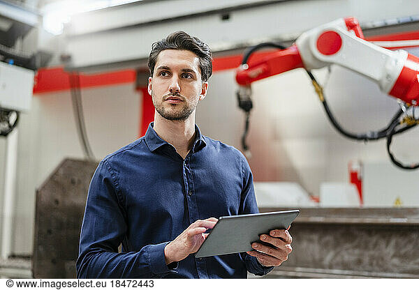 Thoughtful engineer holding tablet PC standing in robot factory
