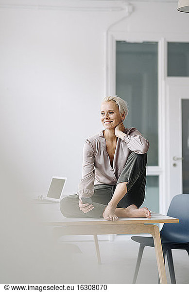 Thoughtful female entrepreneur with smart phone sitting on desk against wall in office