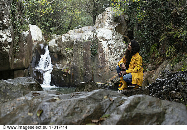 Thoughtful female hiker crouching on rock against waterfall in forest during rainy season