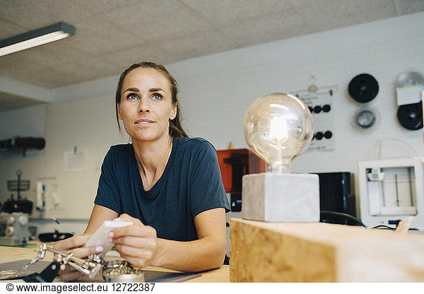 Thoughtful female technician looking away while sitting at workbench in creative office