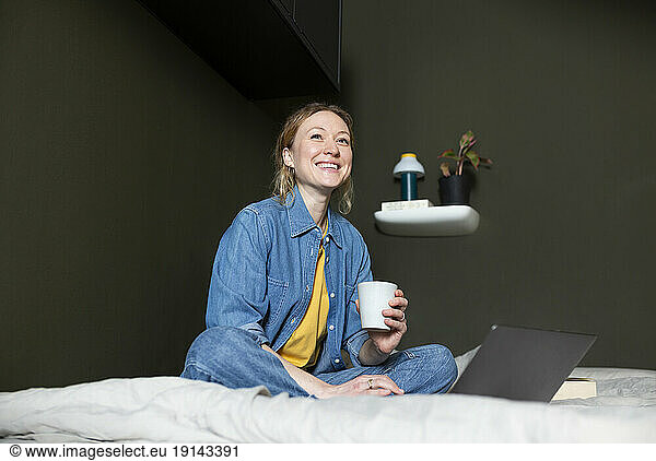 Thoughtful freelancer holding coffee cup with laptop on bed