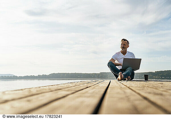 Thoughtful freelancer sitting with laptop on pier over lake