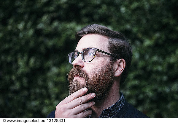 Thoughtful hipster with hand on chin looking away while wearing eyeglasses against hedge