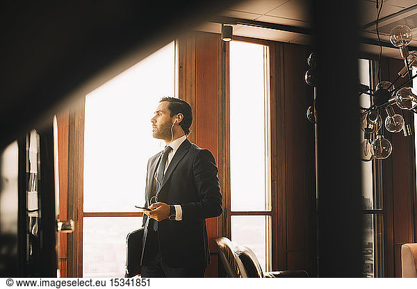 Thoughtful male financial advisor with headphones talking on mobile phone while standing by window at office
