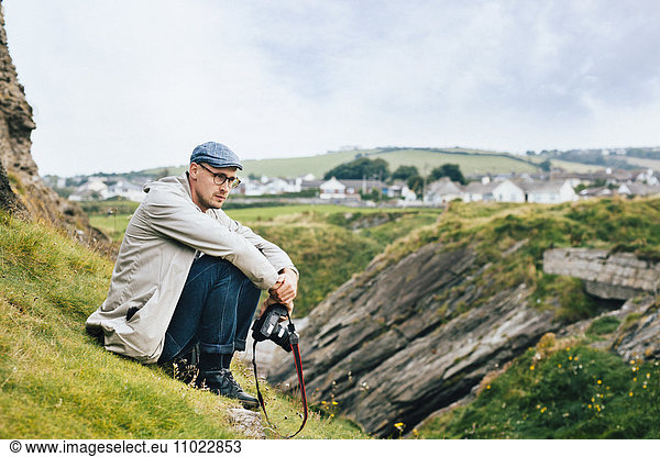 Thoughtful man holding camera while sitting on hill against sky