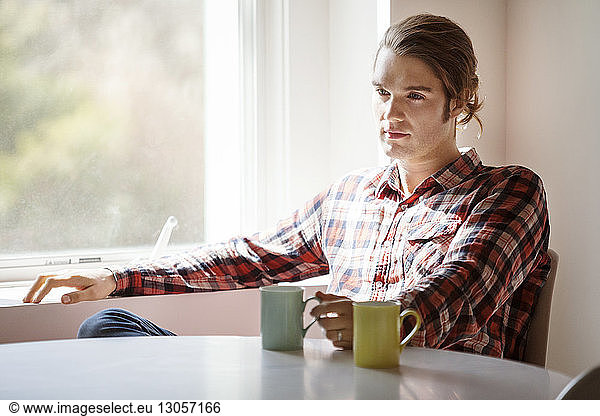 Thoughtful man holding coffee mug while sitting by window at home