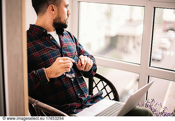 Thoughtful man looking through window working with laptop at home