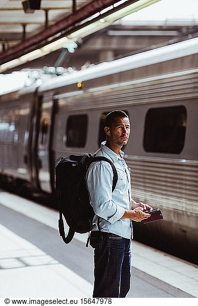 Thoughtful man with backpack holding smart phone while looking at train on platform